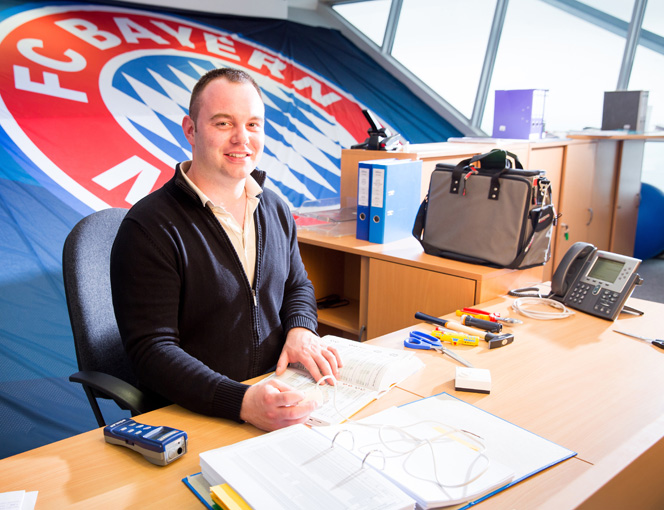 Thomas Sieger sitzt an seinem Schreibtisch, im Hintergrund sieht man ein großes Logo des FC Bayern München.