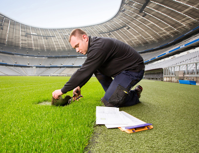 Thomas Sieger kniet am Spielfeldrand und hebt mit der Hand ein Stück Rasen an.